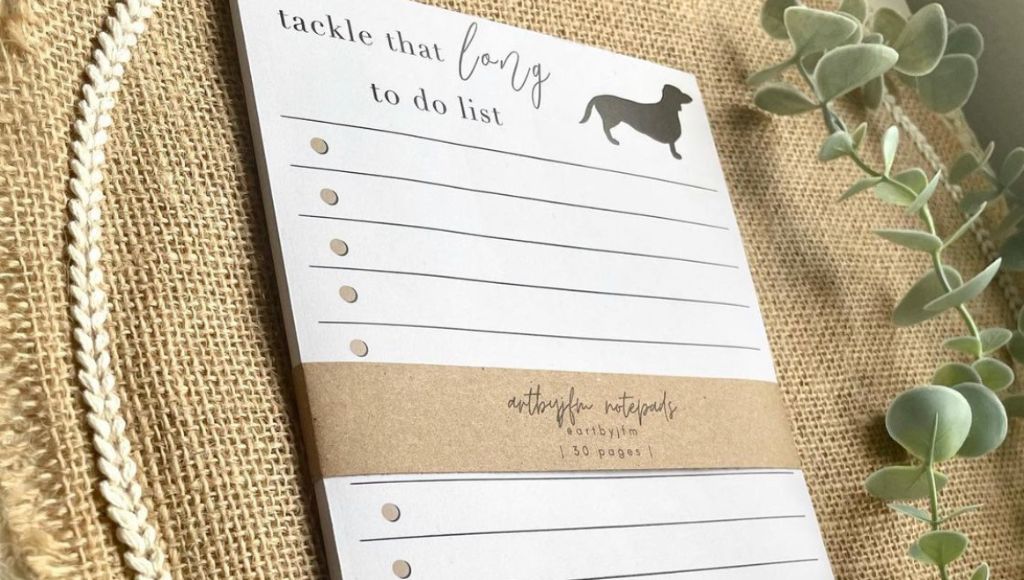 A lined notepad with a silhouette of a dachshund on it and the words "Tackle that long to do list" on the top. The notepad is branded ArtbyJFM.