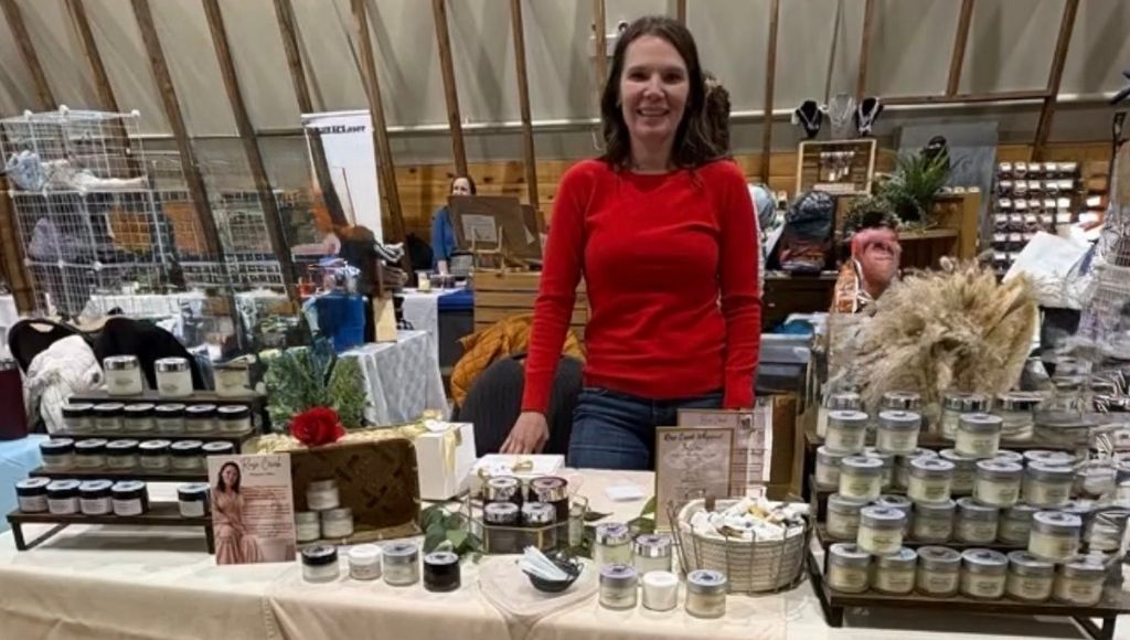 How to Design Info Cards That Sell for You 2 Alice of Rose Creek Whipped Tallow stands at her table at an artisan's market, smiling. Her products, biography card, and pricing card are all visible.