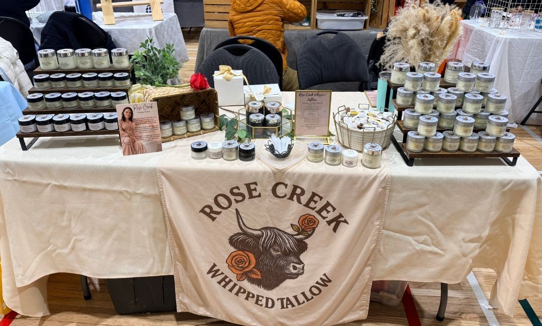 Little Rock Label Printing Alice's booth for her Rose Creek Whipped Tallow company at an artisan's market. An off-white tablecloth is draped over the table with her logo of a bull with roses on it. Her products and biography card are visible.