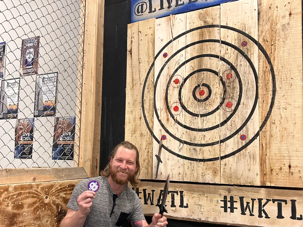 Live Edge Axe Throwing 31 Man holding a branded sticker and axe beside a wooden target at Live Edge Axe, featuring stickers printed at Little Rock Printing.