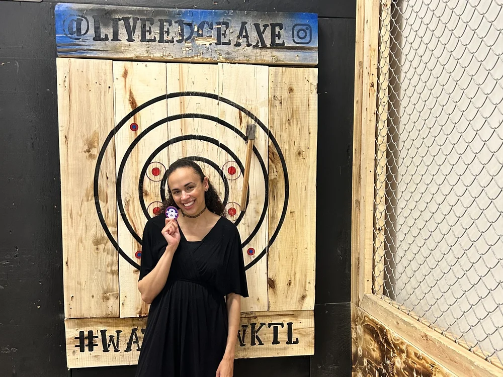 Live Edge Axe Throwing 30 Woman posing with a colourful Live Edge Axe sticker beside a wooden axe-throwing target, printed at Little Rock Printing.