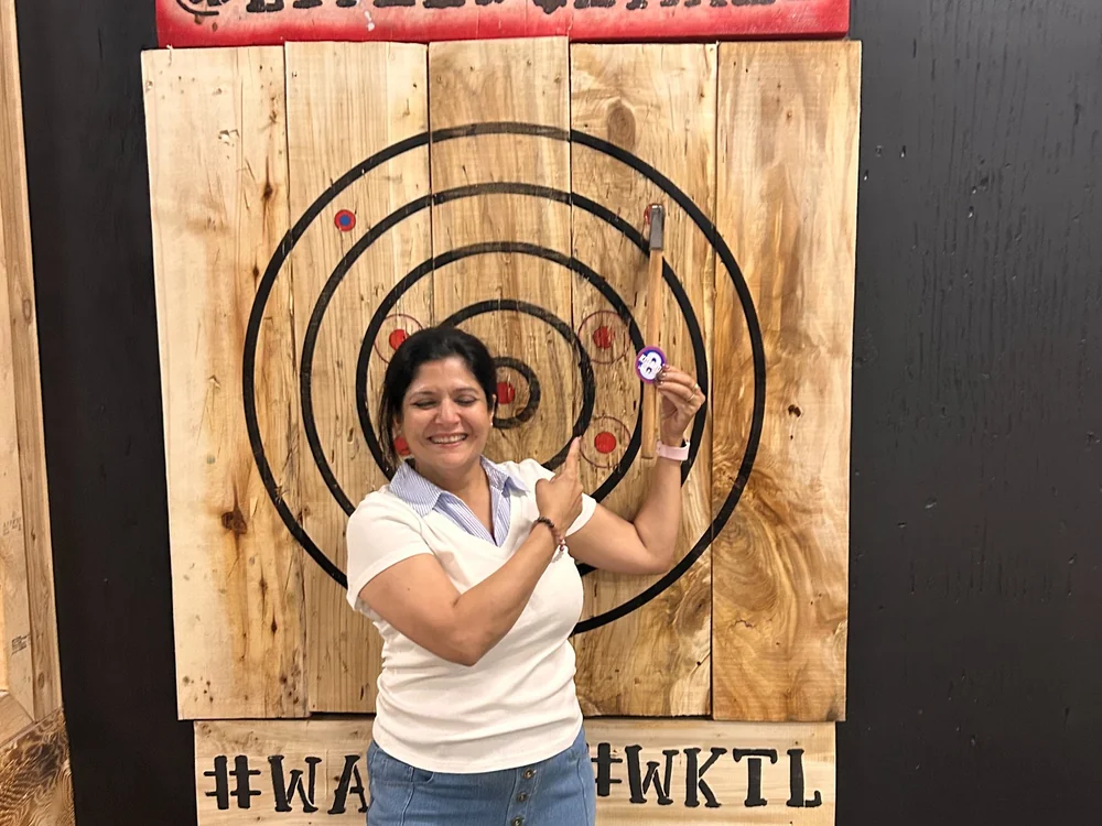 Live Edge Axe Throwing 29 Woman smiling and holding a custom Live Edge Axe sticker in front of an axe-throwing target, printed at Little Rock Printing.