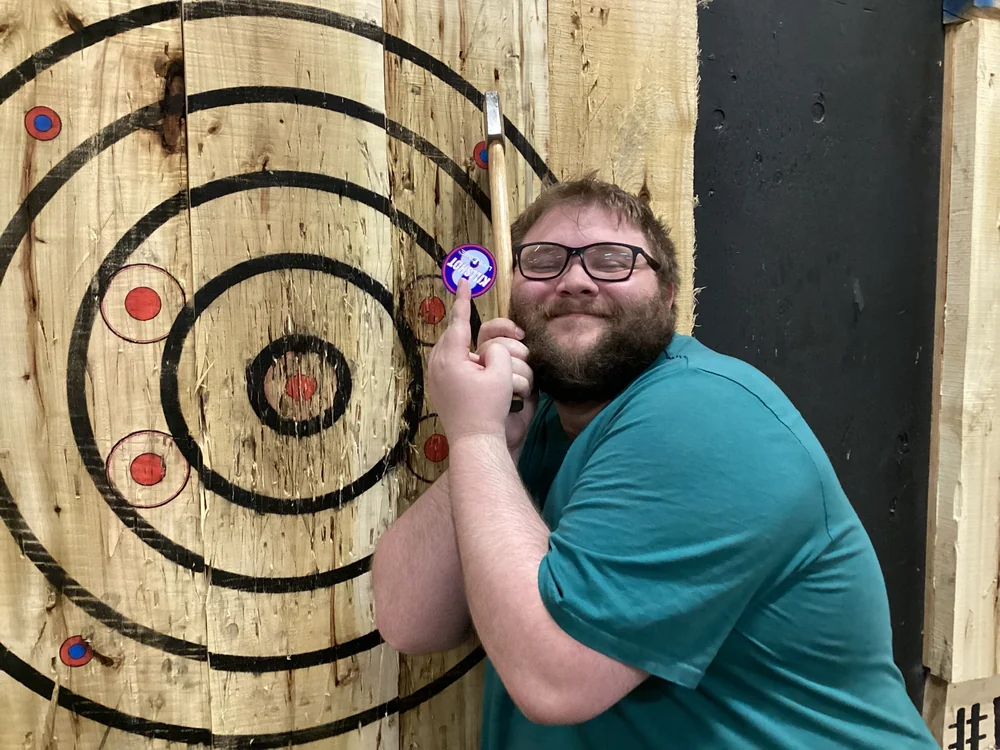 Live Edge Axe Throwing 28 Man holding a branded sticker and axe beside a wooden target at Live Edge Axe, featuring stickers printed at Little Rock Printing.